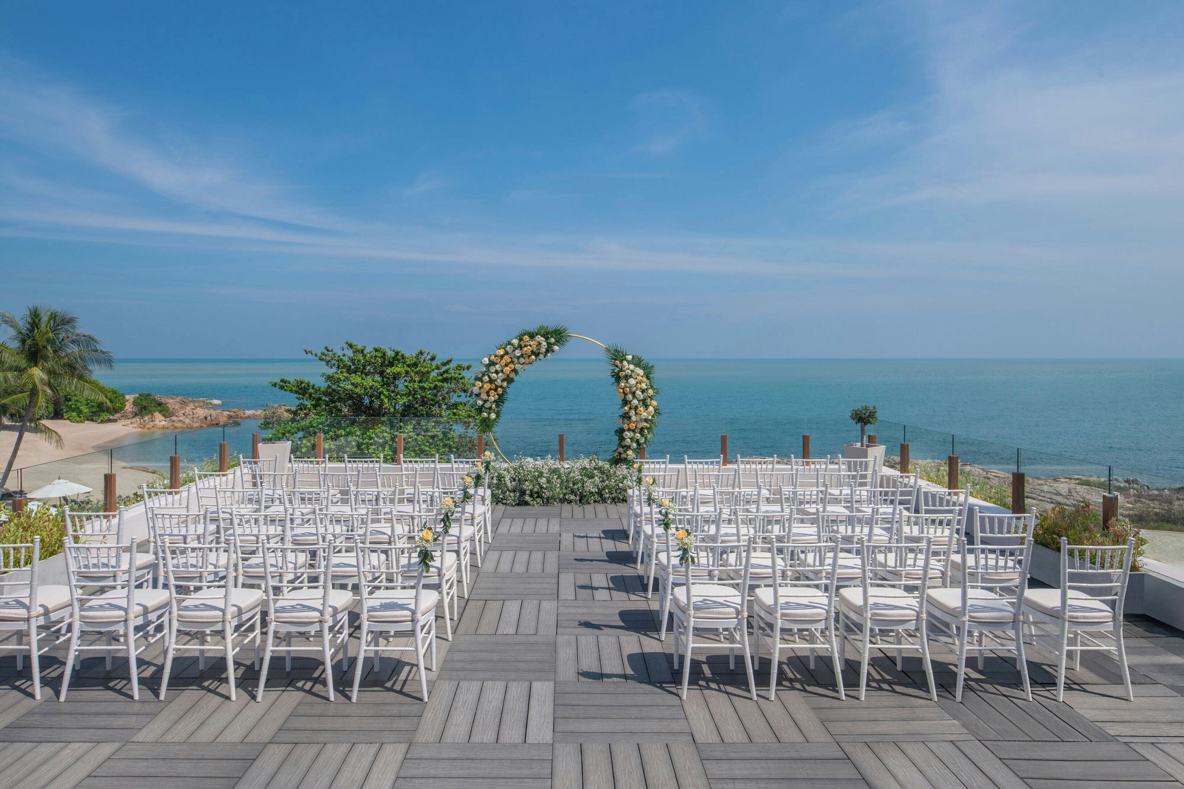 A wedding ceremony setup with white chairs and a floral arch on a wooden deck overlooking the sea.