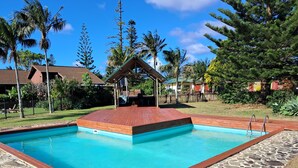 A rectangular swimming pool with a wooden deck and stone border, surrounded by palm trees and a wooden fence. - Fletcher Christian Apartments (Norfolk Island)