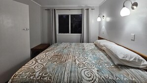 A bedroom with a patterned bedspread, a ceiling fan, a window with curtains, and wall-mounted lights. - Fletcher Christian Apartments (Norfolk Island)