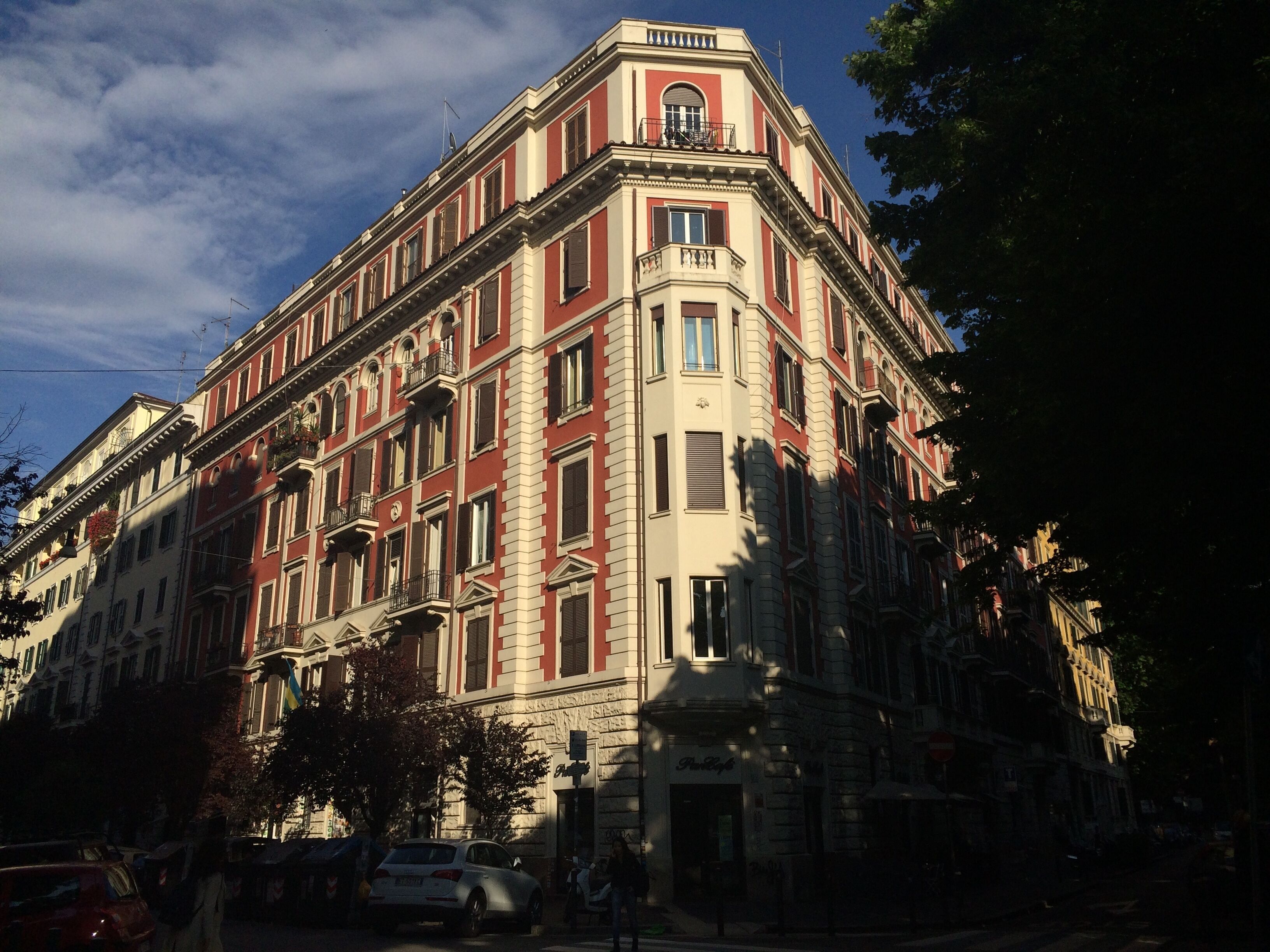 Un immeuble à plusieurs étages, de style architectural classique, avec une entrée d’angle, de grandes fenêtres et un balcon aux étages supérieurs.