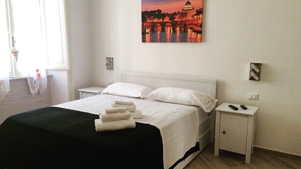 Un lit bien fait, avec des draps blancs et un jeté noir, une table de chevet avec une télécommande, et un tableau représentant une vue de la ville au crépuscule, accroché au mur.
