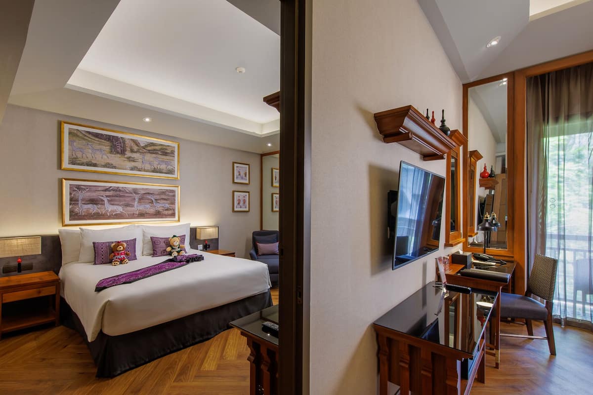 A hotel room with a large bed, a desk, a chair, and a TV. There are framed pictures on the wall and a mirror above the desk.