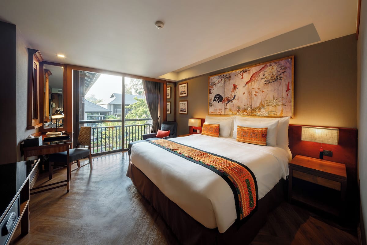 A hotel room with a large bed, a desk, a chair, a balcony view, and a painting on the wall.