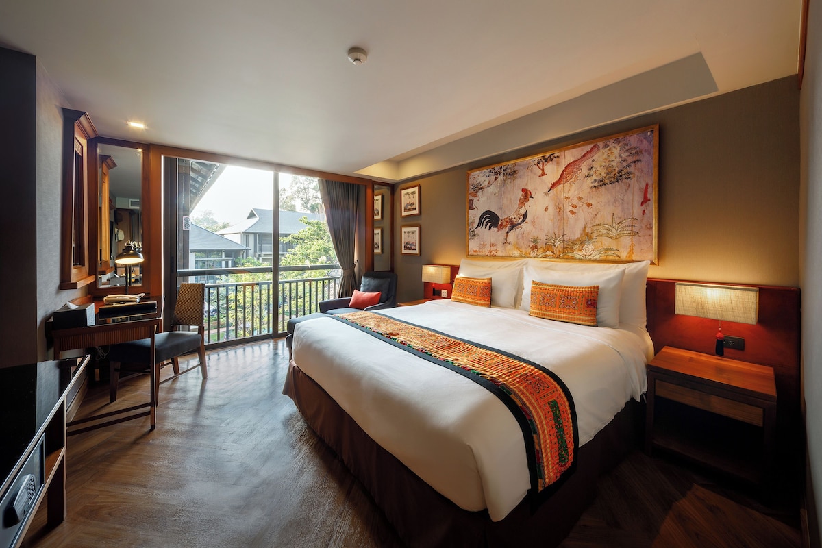 A hotel room with a large bed, a desk, a chair, a balcony view, and a painting on the wall.