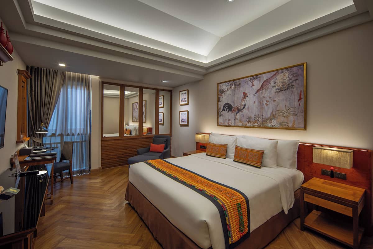 A hotel room with a large bed, a desk, and a chair. There is a painting on the wall and a window with curtains.