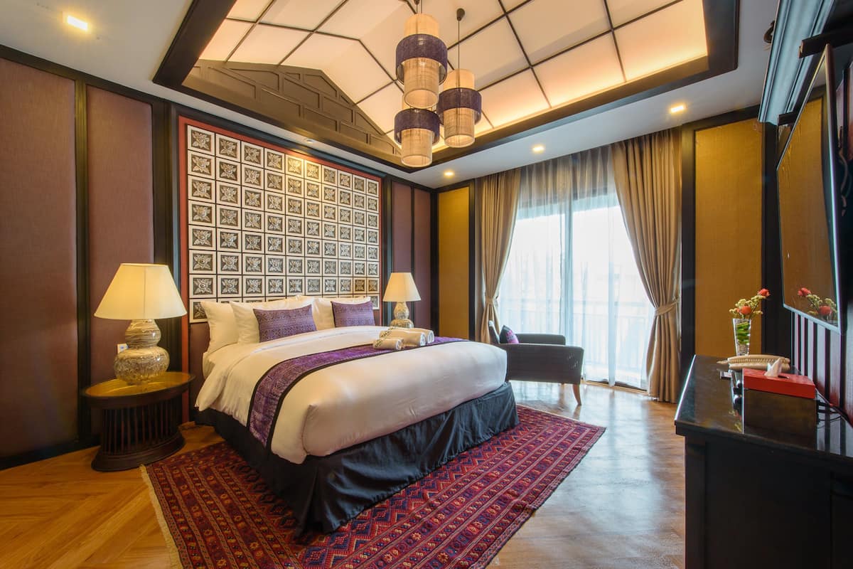 A hotel room with a large bed, two bedside tables with lamps, a decorative wall panel, a desk with a vase, and a large window with curtains.