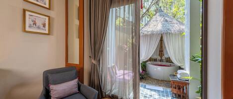 A room with a grey sofa, a large window with sheer curtains, and a view of an outdoor area with a bathtub and a thatched structure.