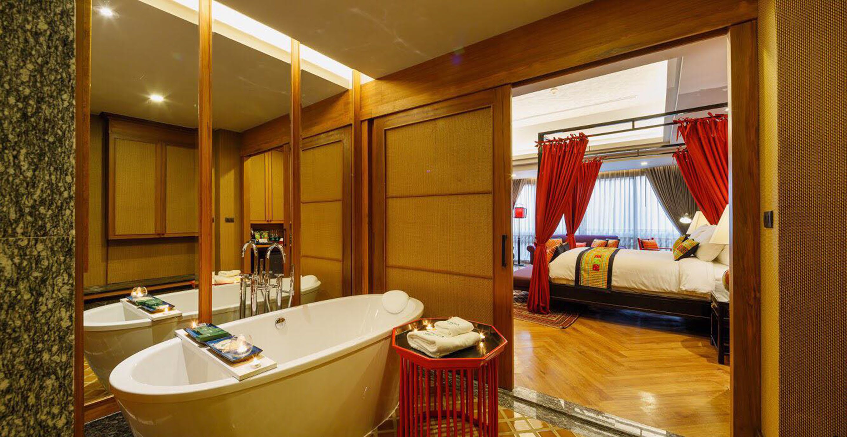Un baño de hotel con una bañera grande, zona de ducha separada y un dormitorio con cama, cortinas y ventana.