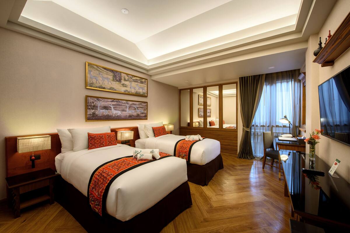 A hotel room with two beds, a desk, a chair, a TV, and artwork on the walls.