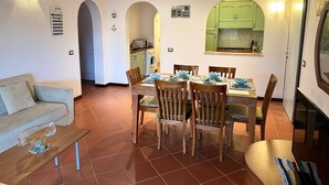 A dining area with a table set for four, surrounded by wooden chairs. There is a sofa and a coffee table with a decorative bowl. A kitchen is visible through an archway, with a clock and a 'LOVE' sign on the wall.