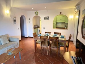 Uma sala de jantar com uma mesa posta para quatro pessoas, rodeada por cadeiras de madeira. Há um sofá e uma mesa de centro com uma tigela decorativa. Uma cozinha é visível através de um arco, com um relógio e uma placa com a palavra 'LOVE' na parede.