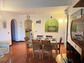A dining area with a table set for four, surrounded by wooden chairs. There is a kitchen visible through an archway, and a clock on the wall. - SPECIAL EDITION 2020 - BEAUTIFUL EXCLUSIVE VILLA WITH PRIVATE VILLAGE POOL (Stintino)