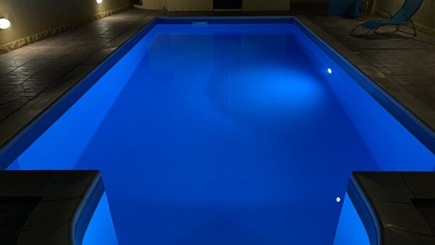 A rectangular swimming pool with a blue illuminated interior, surrounded by stone flooring and a chair in the background.