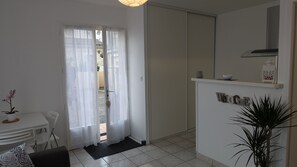 Une pièce moderne et bien éclairée, dotée d’une porte coulissante donnant sur un espace extérieur, d’une petite table à manger avec des chaises, d’une plante en pot et d’une pancarte de bienvenue accrochée au mur.