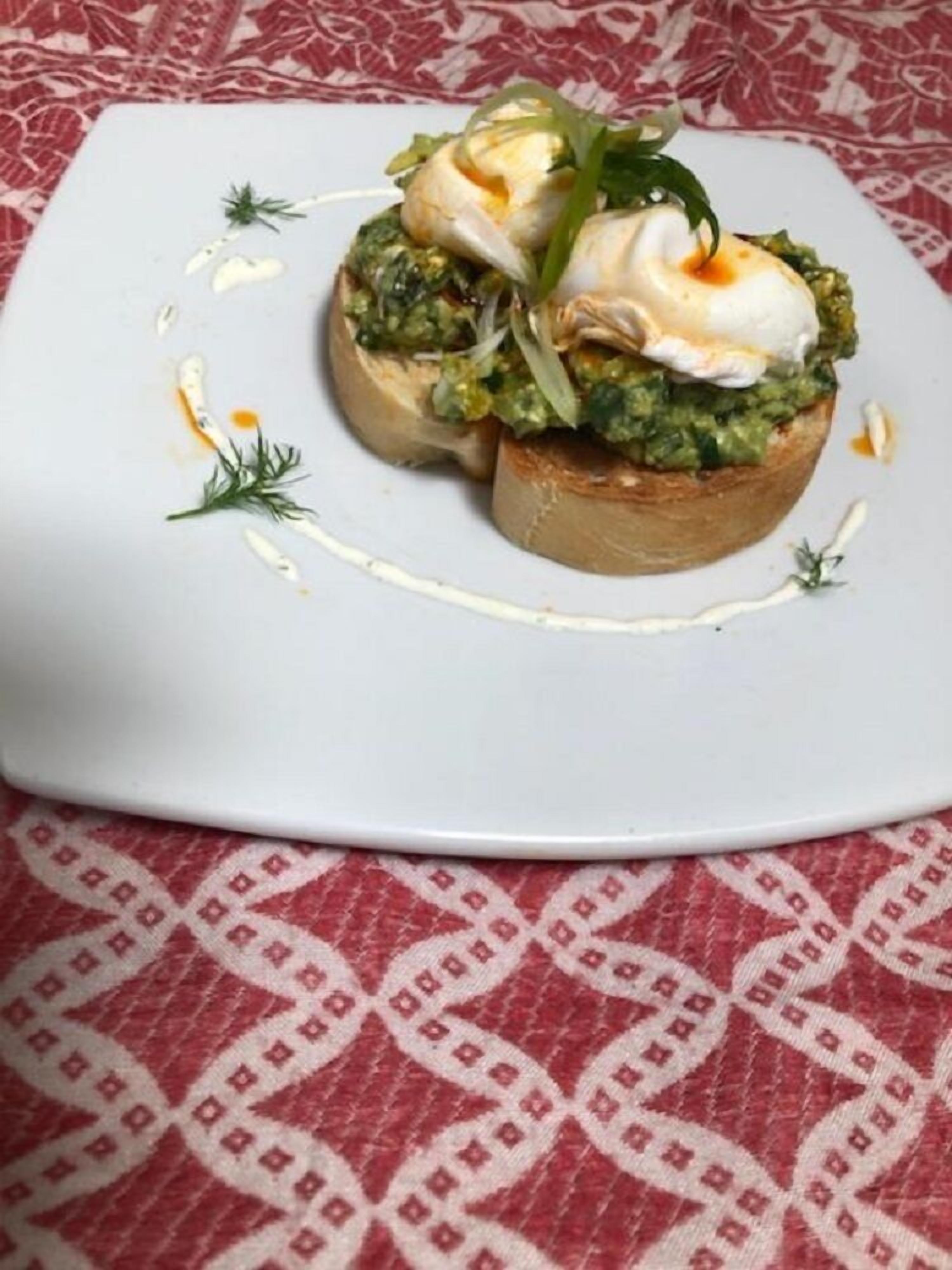 A square white plate with a dish of bruschetta topped with a poached egg, pesto, and greens, on a patterned tablecloth.