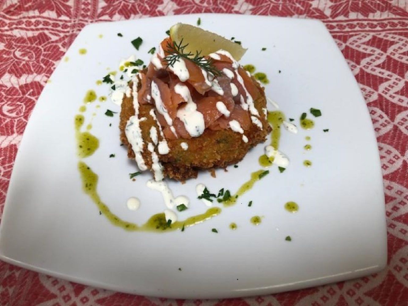 Un plato blanco cuadrado con un trozo de comida cubierto con una rodaja de limón y hierbas, rociado con salsa blanca y aceite verde, sobre un mantel estampado.