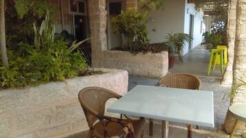 An outdoor seating area with wicker chairs and a table, surrounded by greenery and stone walls.
