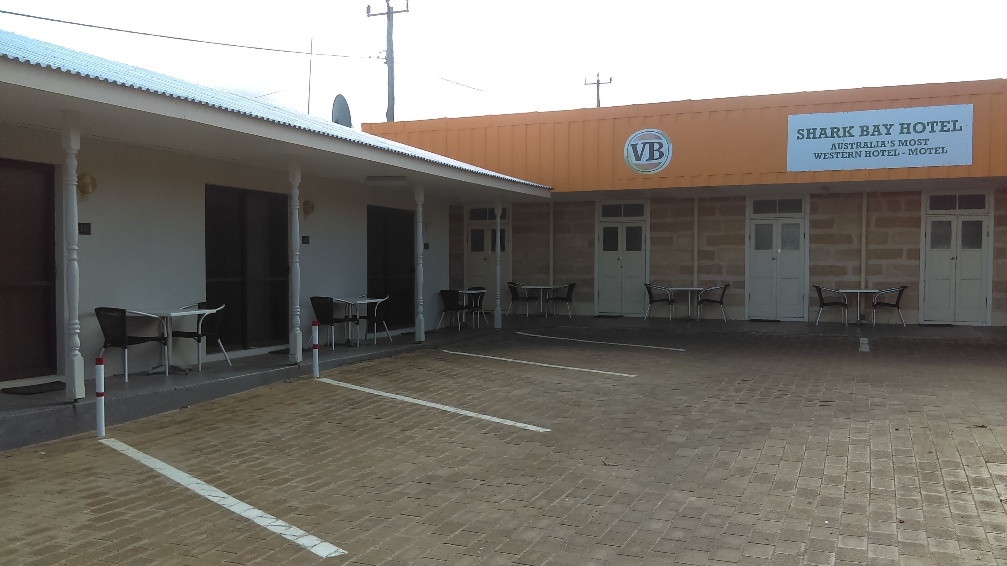 A hotel with a covered outdoor seating area, multiple tables and chairs, and a sign indicating it is Shark Bay Hotel.
