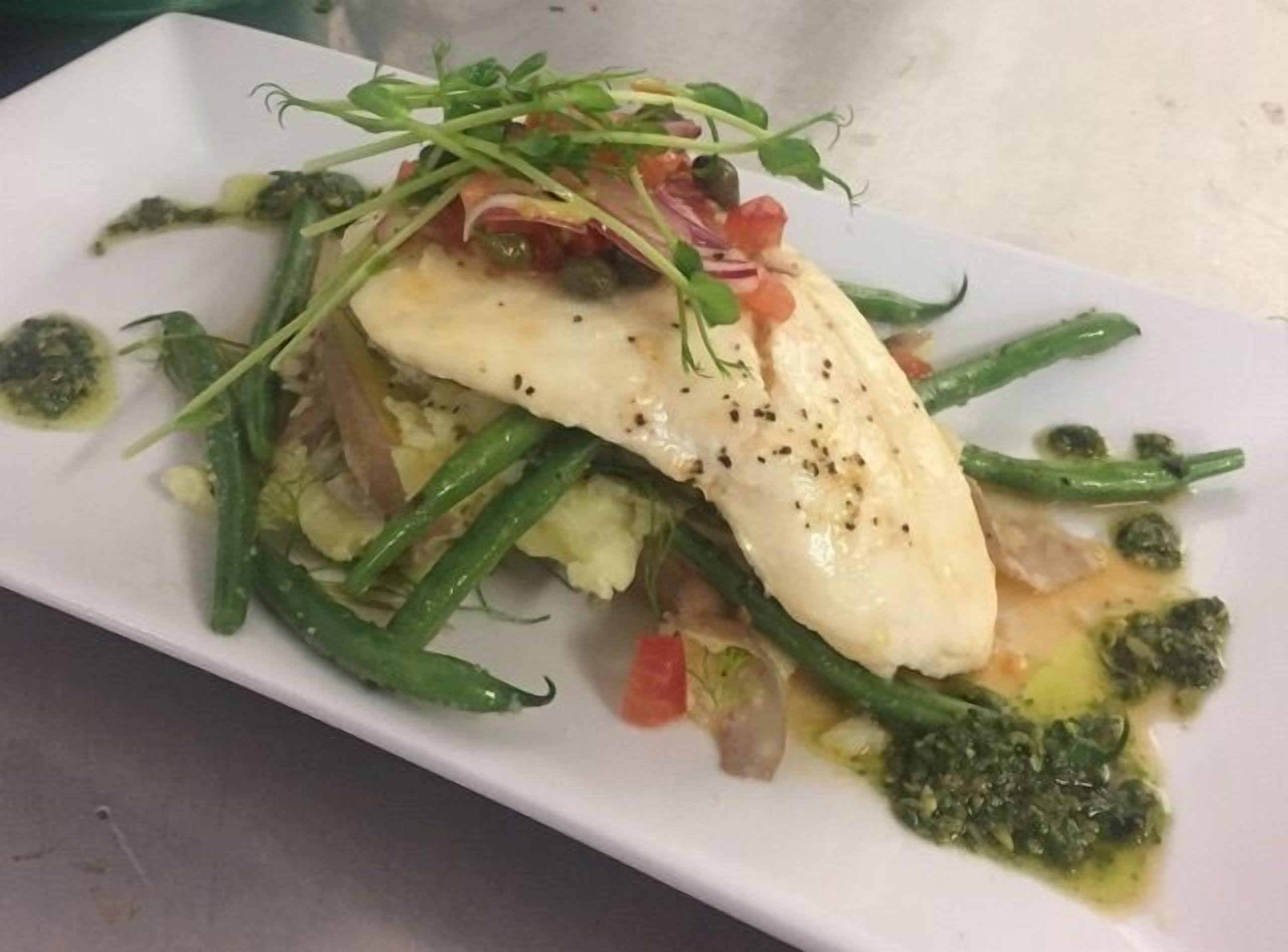 A white rectangular plate with a piece of white fish topped with green herbs, green beans, and a green sauce.