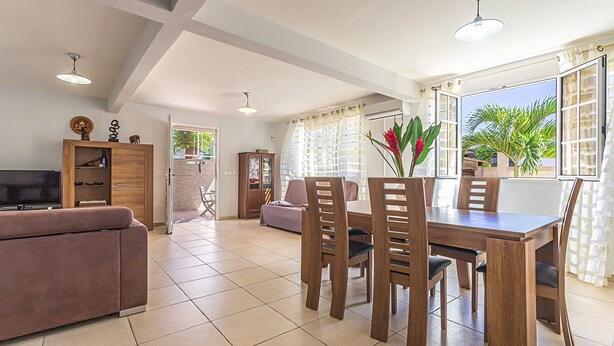 A spacious living area with a sofa, dining table, and chairs, a television, and a large window with curtains. - Bottom of villa with pool (le Moule)