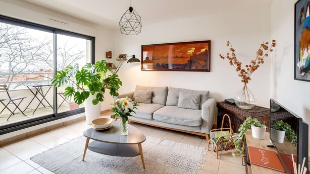 A modern living room with a grey sofa, a coffee table, and a large window offering a view of the city. - Votre Escale - Le Petit Montmartre (Niort)
