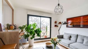 A modern living room with a grey sofa, a coffee table, a large window with a view of trees, and a cactus plant. - Votre Escale - Le Petit Montmartre (Niort)
