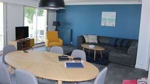 Un salón moderno con una mesa redonda de madera, sillas grises y amarillas, un sofá gris, una pequeña mesa redonda de centro y un televisor sobre un mueble de madera.