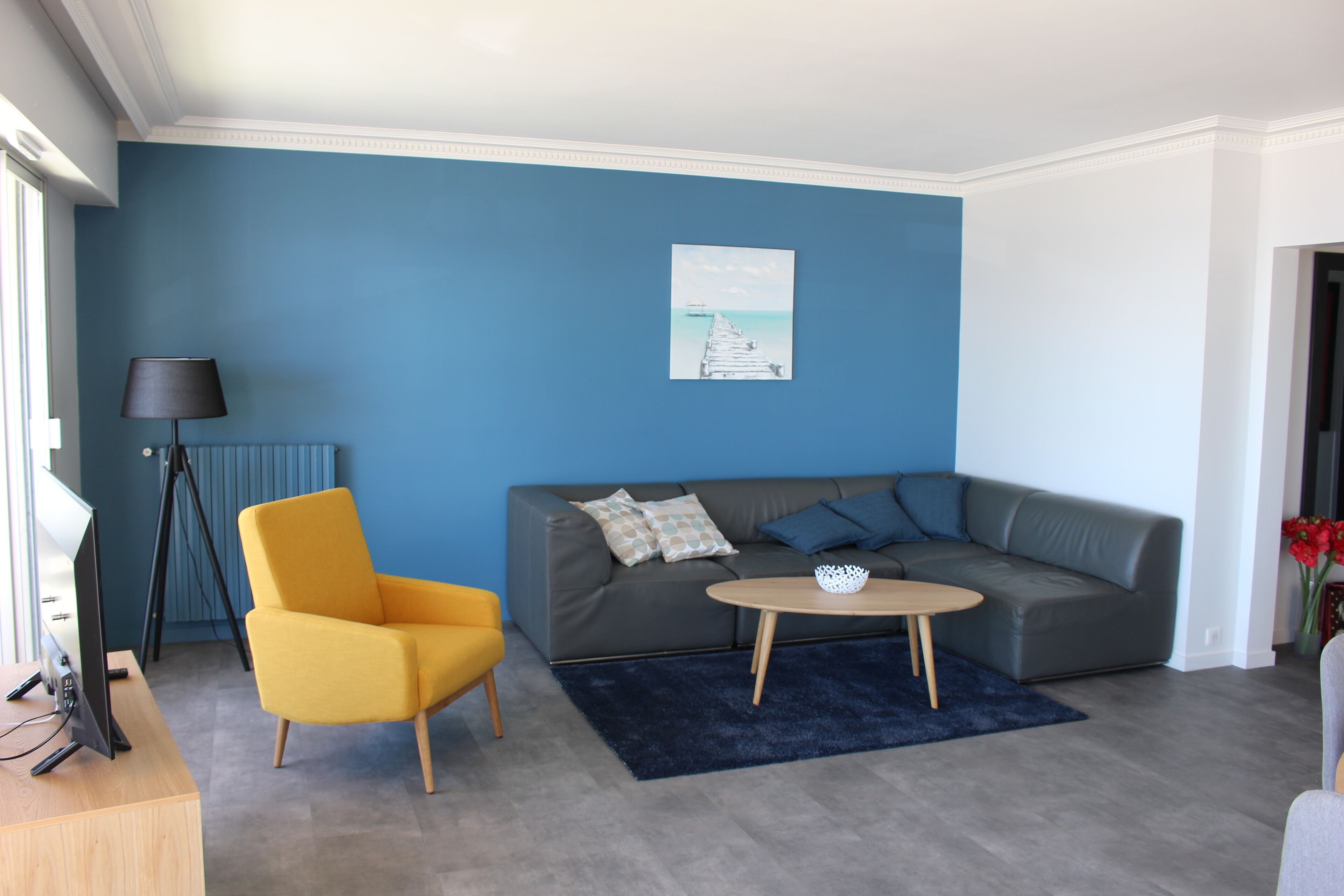 Un salon moderne avec un canapé gris, un fauteuil jaune, une table basse ronde et un mur d’accent bleu.