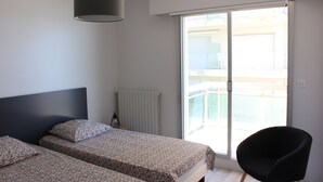 Un dormitorio con dos camas, una silla negra, un radiador y una ventana con vista a un balcón.