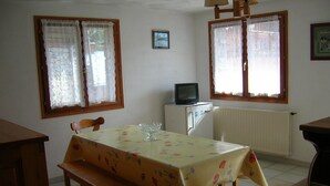 A dining area with a table covered in a floral tablecloth, a bench, a television on a small cabinet, and lace curtains on the windows. -  BEAUTIFUL GITE 4 PEOPLE  (Bozel)