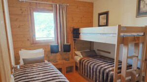 Ein schlicht eingerichtetes Zimmer im Blockhausstil mit zwei Etagenbetten, einem Fenster mit Vorhängen, einem Nachttisch mit Lampe sowie einem gerahmten Bild an der Wand.