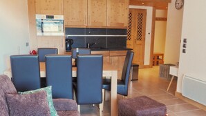 Ein modernes Wohnzimmer mit einer Couch, Sesseln und einem Couchtisch. Im Hintergrund ist eine Küche mit Holzschränken und einem Spülbecken zu sehen.