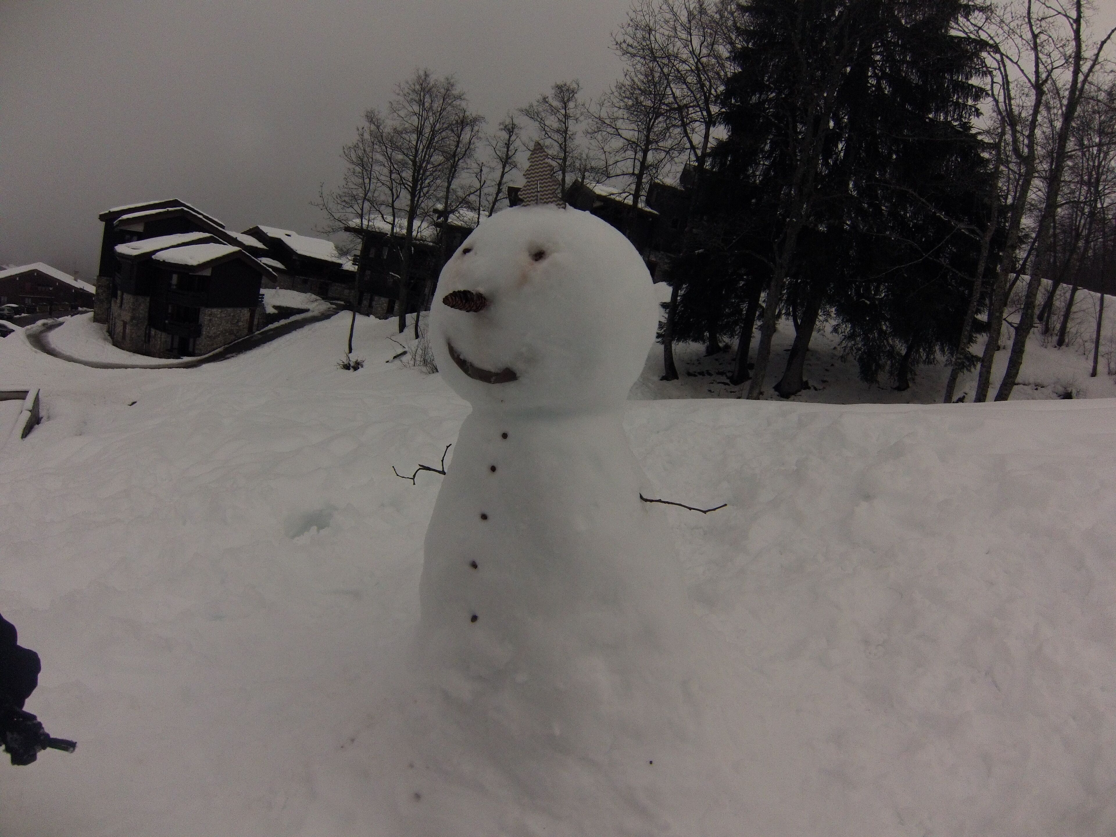 Un bonhomme de neige dans un décor hivernal, avec des arbres sans feuilles et un bâtiment en arrière-plan.