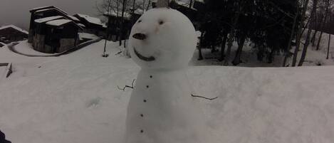 Un bonhomme de neige dans un décor hivernal, avec des arbres sans feuilles et un bâtiment en arrière-plan.