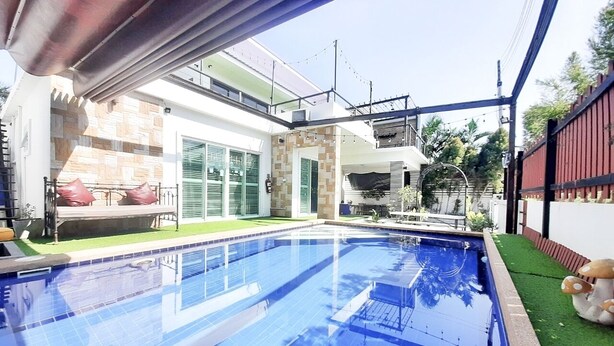 A swimming pool with a tiled deck, a covered seating area, and a view of a house with a balcony and outdoor furniture. - Baan Bua Sawhan Pool Villa Hua Hin (Hua Hin)