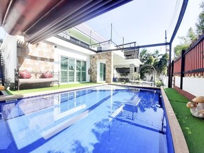 A swimming pool with a tiled deck, a covered seating area, and a view of a house with a balcony and outdoor furniture. - Baan Bua Sawhan Pool Villa Hua Hin (Hua Hin)