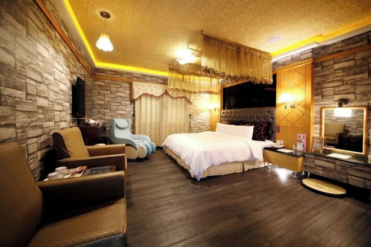 Habitación de hotel con pared de piedra, una cama grande, zona de estar con una silla, una mesita y un espejo.