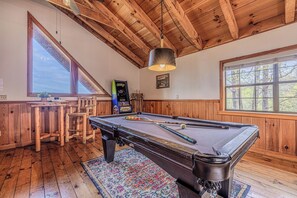 Una mesa de billar en una habitación con paredes y techo de madera, una zona de bar y ventanas amplias.
