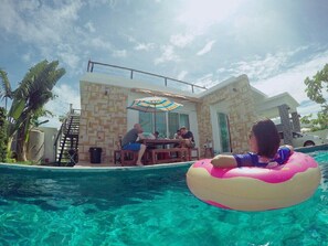 A pool area with a stone building, a table with people, and a person in a pink inflatable ring. - Baan Khunnatham Pool Villa Hua Hin (Hua Hin)