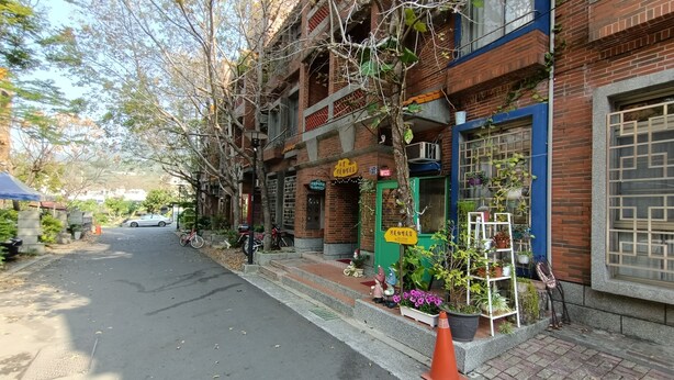A street with a row of brick buildings, some with visible signage. There are bicycles parked outside, and a few potted plants are placed on the sidewalk. - Shuili Heti (Shuili)