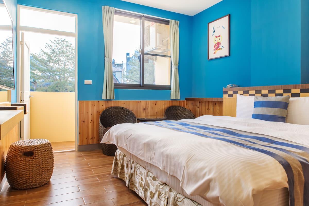A bedroom with a large bed, a window with curtains, a wooden floor, and a view of the outside.