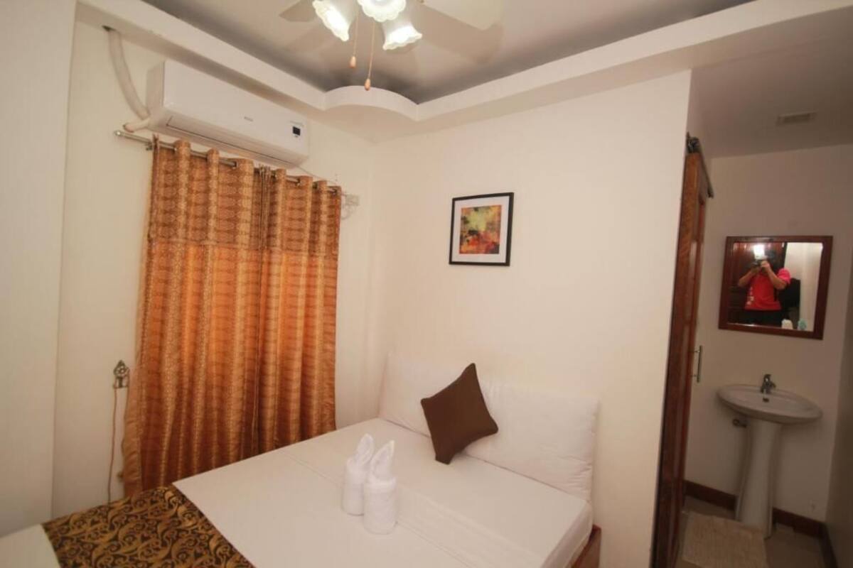 A hotel room with a bed, a pillow, a framed picture on the wall, an air conditioning unit, a curtain, and a sink.