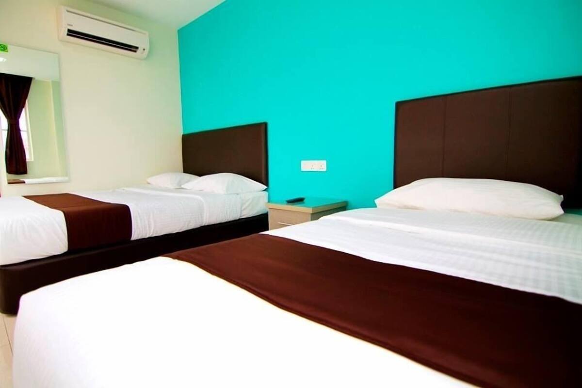 Habitación de hotel con dos camas, cada una con cabecero marrón y ropa de cama blanca. Hay una ventana con cortinas, un equipo de aire acondicionado y una mesita de noche pequeña con una lámpara.