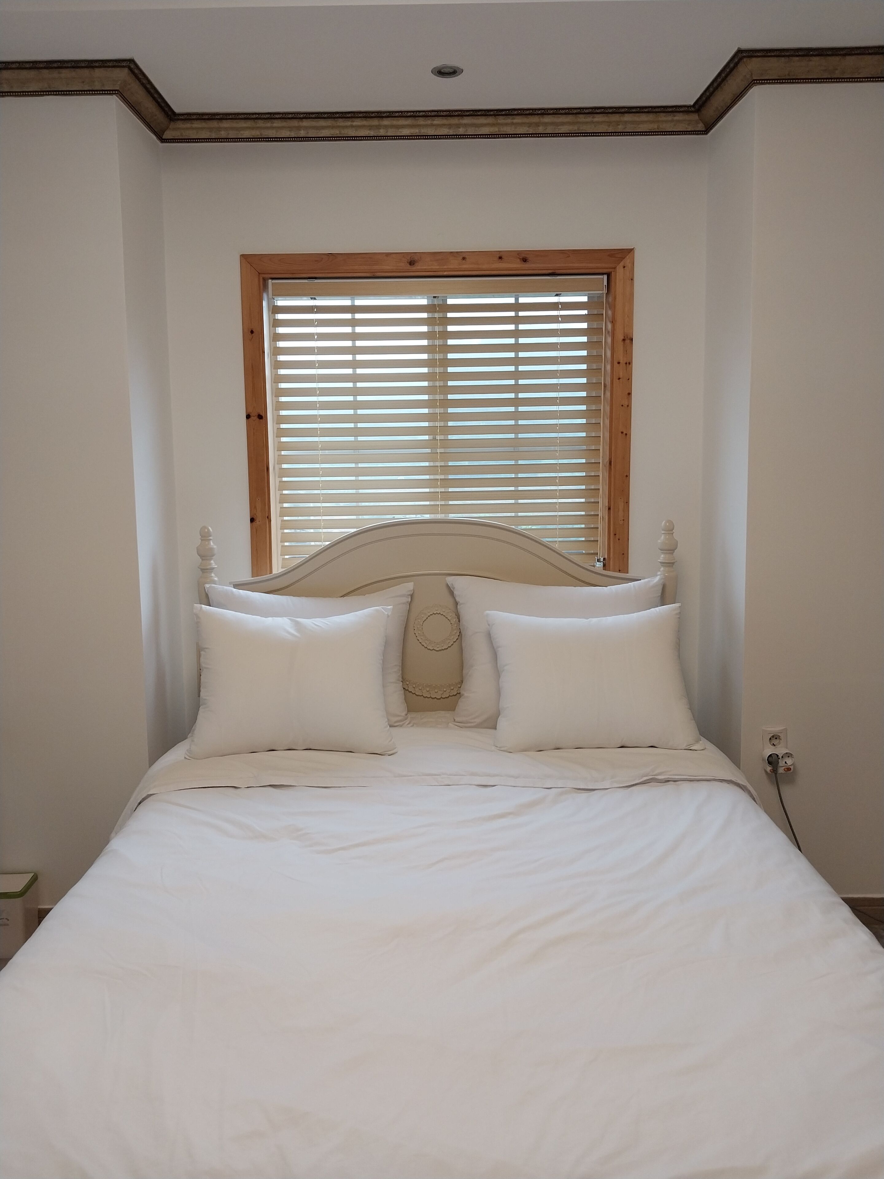 Una cama bien hecha con ropa de cama blanca y un cabecero de madera, una ventana con persianas y un enchufe de pared.