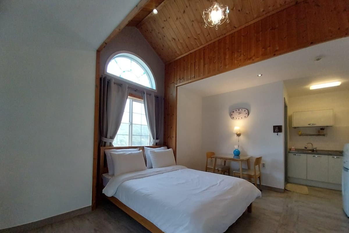 Un dormitorio con cama, una mesita, una silla, un reloj y una ventana con cortinas.