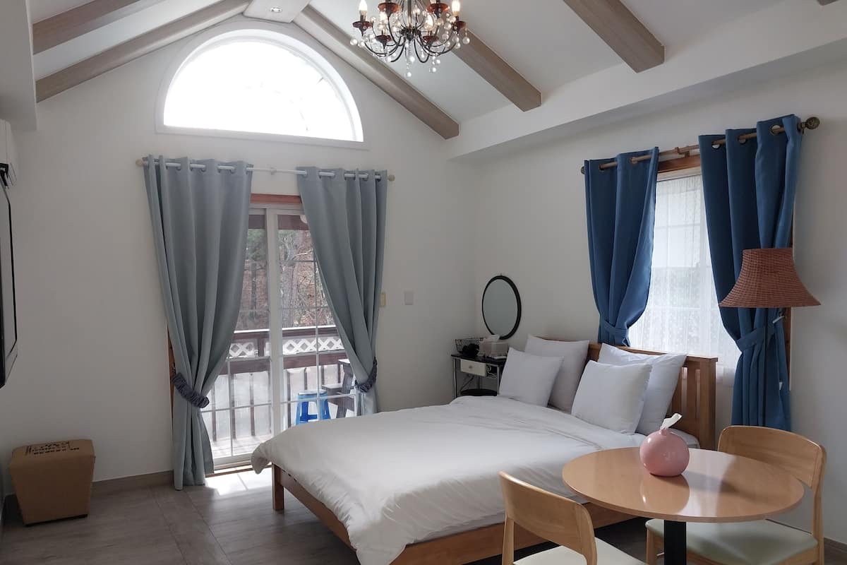 Un dormitorio con cama, una mesa redonda de madera con un florero rosa, una silla de madera, un taburete pequeño, una mesita de noche con espejo y una ventana con cortinas.