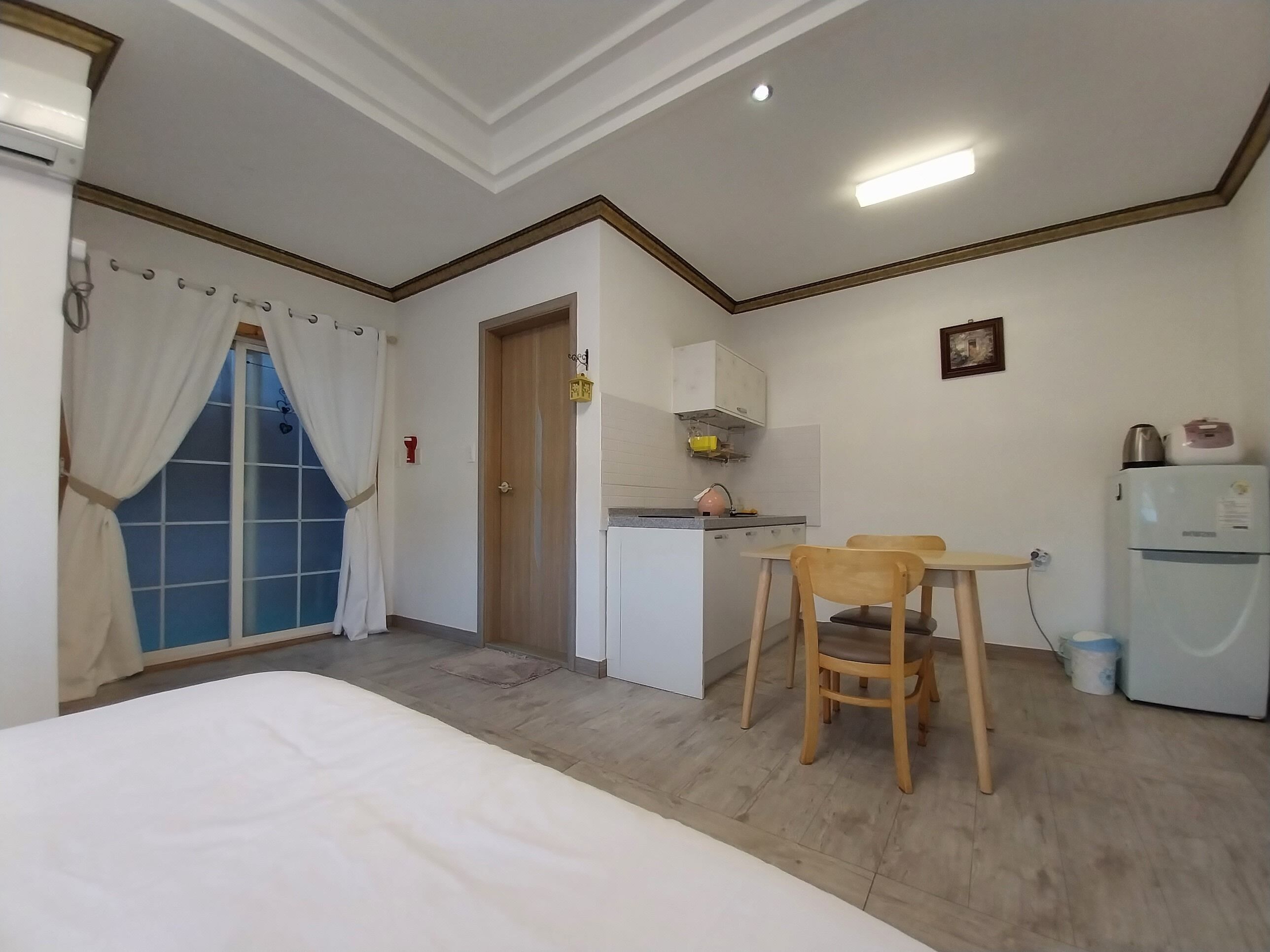 Habitación compacta con cama, una pequeña kitchenette, una mesa de comedor con sillas y un ventanal con cortinas.