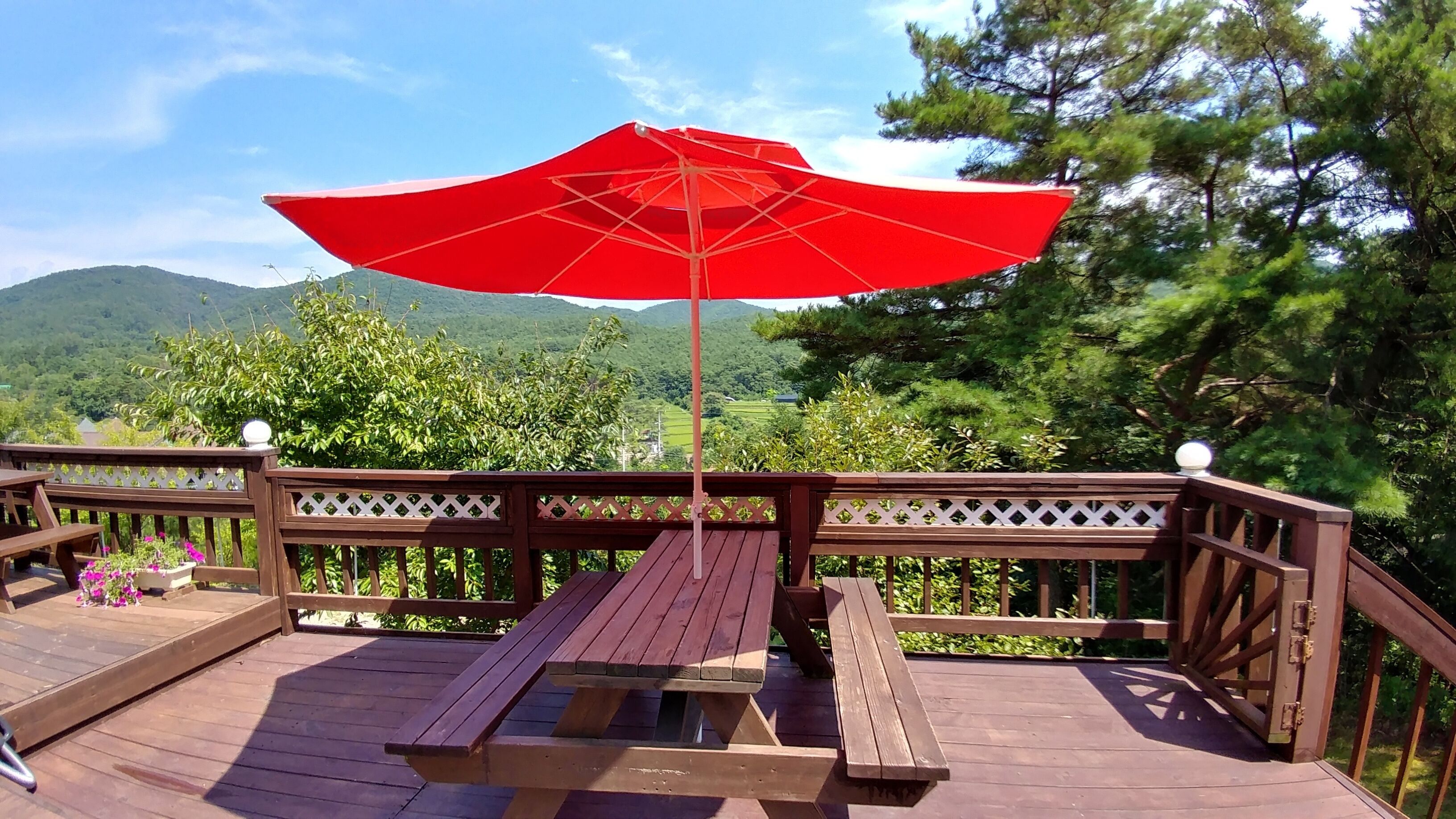 Une terrasse en bois avec un parasol rouge, une table de pique-nique et des bancs.