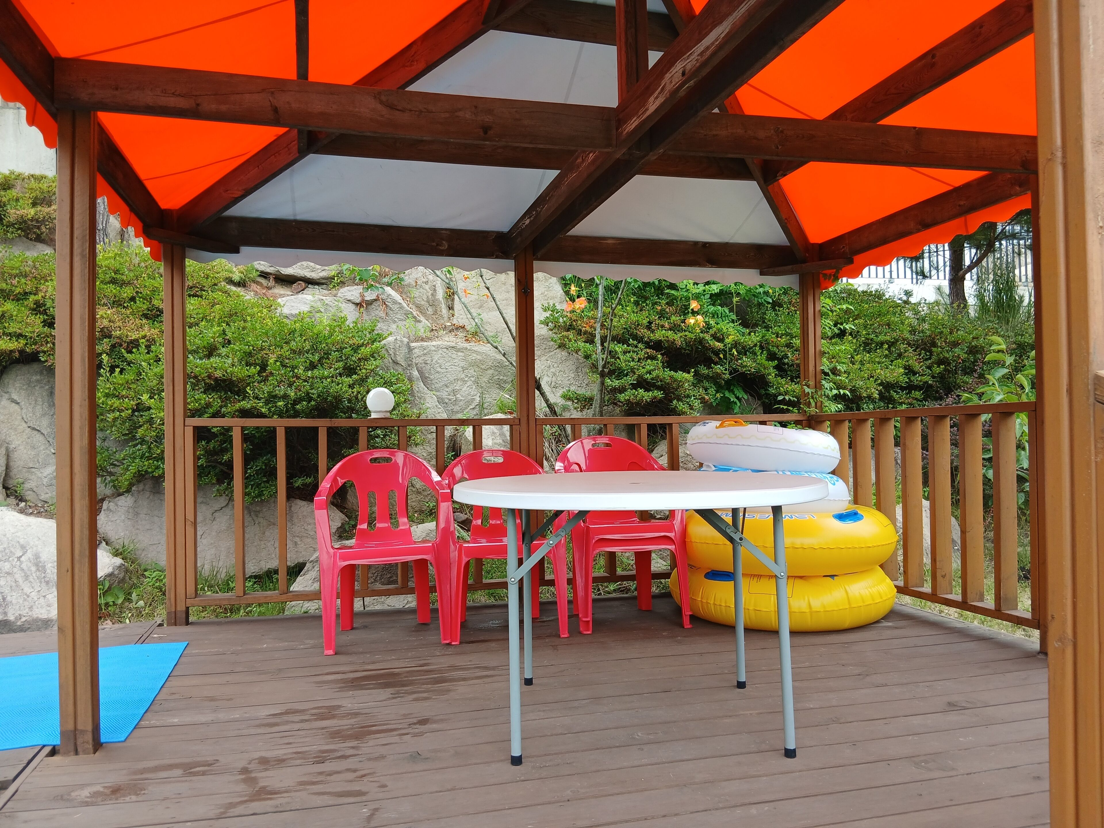 A covered outdoor seating area with wooden beams and orange canopies, featuring red plastic chairs and a white table.