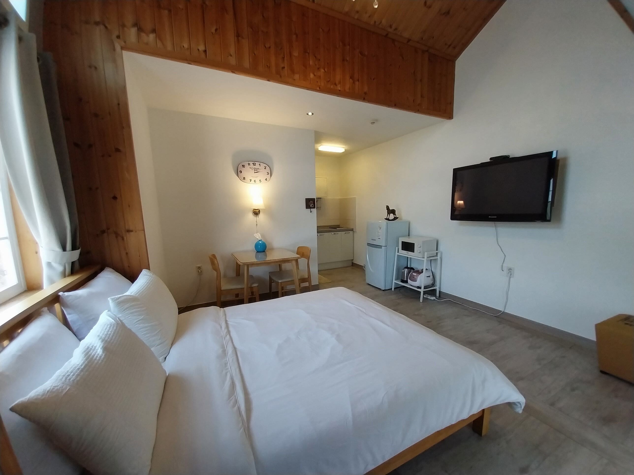 A bedroom with a bed, a small table with chairs, a wall-mounted TV, and a kitchenette in the background.